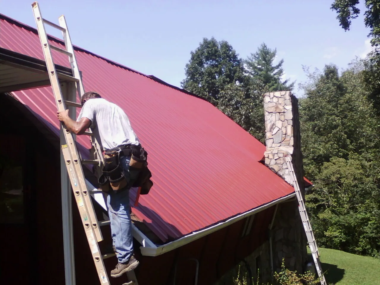 Roofer on a ladder beside a red metal roof and stone chimney — Soffit & Fascia Repair by Roofing Brothers in Moscow